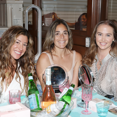 Three women sitting together at a table with drinks and makeup mirrors.